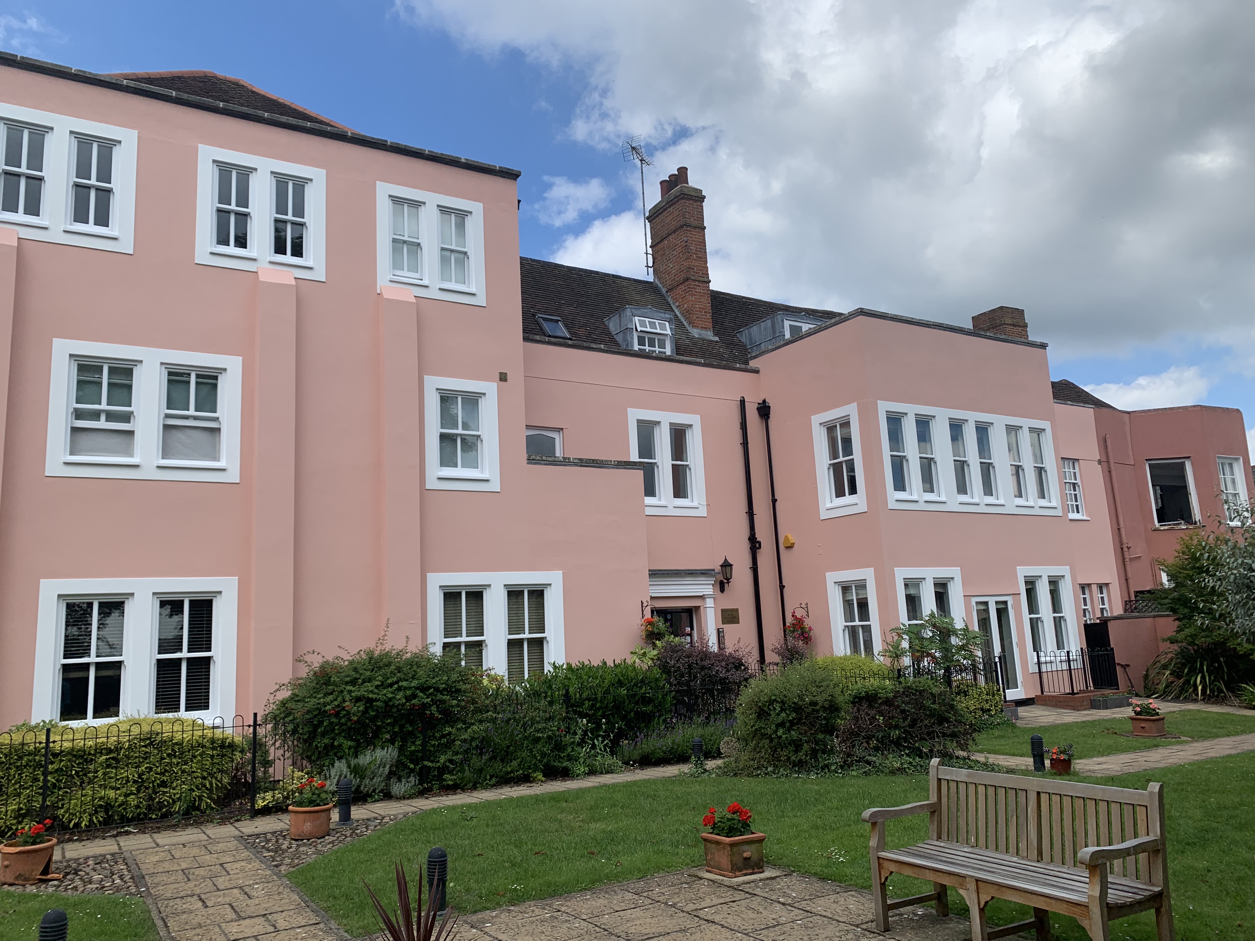 A pink residential building with white trim, featuring multiple windows and a well-maintained garden with flowers and a bench in the foreground.