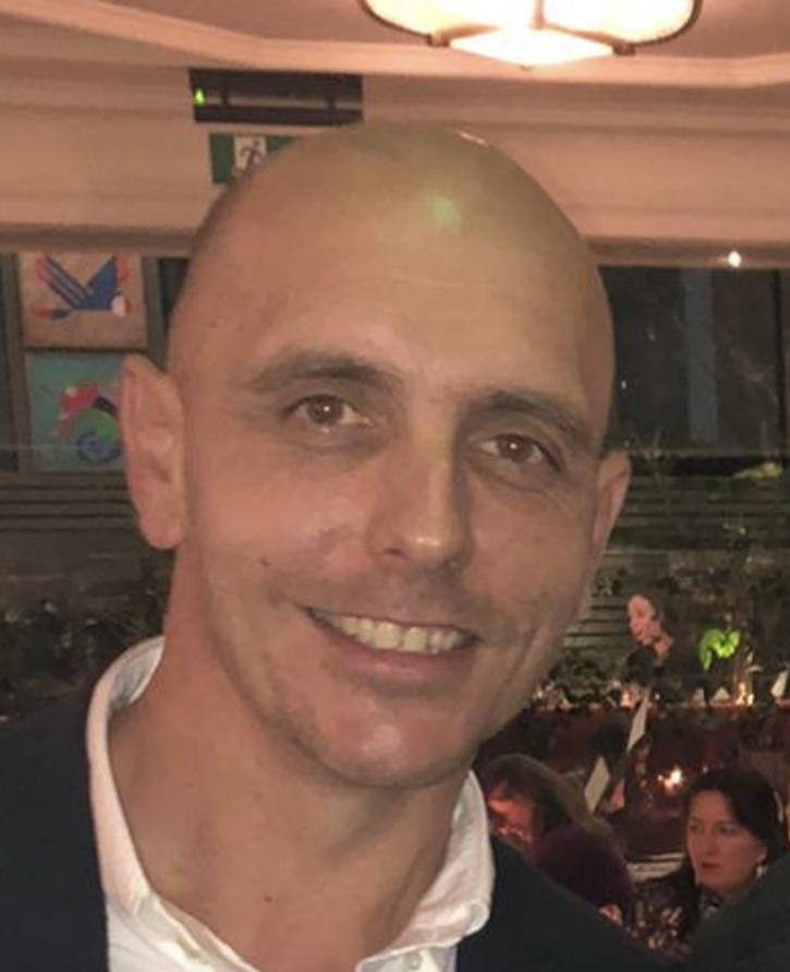 A man with a shaved head and a friendly smile, dressed in a dark jacket with a white shirt, standing in a social setting.