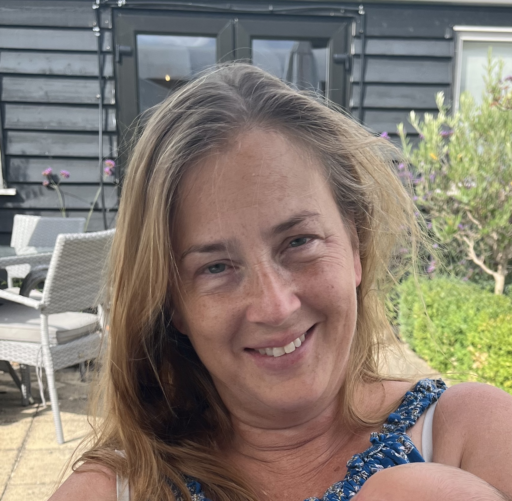 A woman with long, wavy hair smiles at the camera, sitting outdoors in a garden-like setting with green foliage and patio furniture in the background.